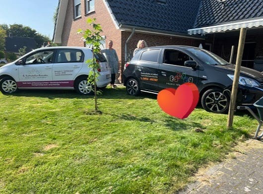 Zwei Autos stehen vor einem Haus mit einem neu gepflanzten Baum auf dem Rasen, zwei Personen stehen zwischen den Fahrzeugen